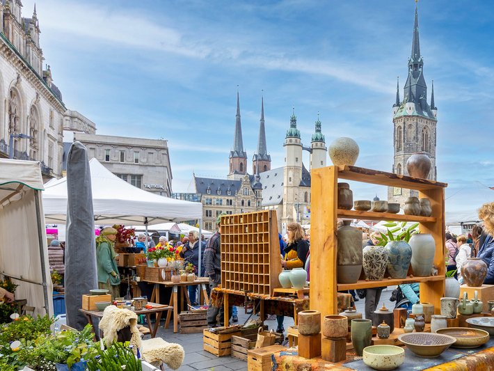 Buntes Marktstand-Bild mit handgefertigten Keramikprodukten wie Vasen, Töpfen und Schalen auf Regalen und Tischen. Im Hintergrund historische Gebäude mit Türmen und Zeltstrukturen auf einem belebten Platz.