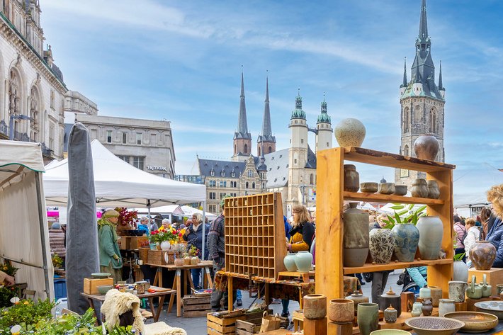 Buntes Marktstand-Bild mit handgefertigten Keramikprodukten wie Vasen, Töpfen und Schalen auf Regalen und Tischen. Im Hintergrund historische Gebäude mit Türmen und Zeltstrukturen auf einem belebten Platz.
