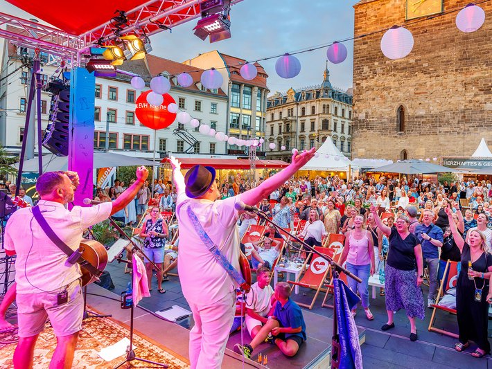 Auf einem bunten Straßenfest tanzen zwei Personen in pinken Anzügen mit aufwendigen Pailletten vor einem Publikum, während im Hintergrund Luftballons in verschiedenen Farben schweben und eine Bühne mit Musikinstrumenten zu sehen ist.