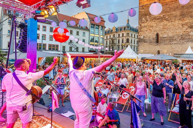 Thomas Ziegler, Stadt Halle (Saale) Auf einem bunten Straßenfest tanzen zwei Personen in pinken Anzügen mit aufwendigen Pailletten vor einem Publikum, während im Hintergrund Luftballons in verschiedenen Farben schweben und eine Bühne mit Musikinstrumenten zu sehen ist.