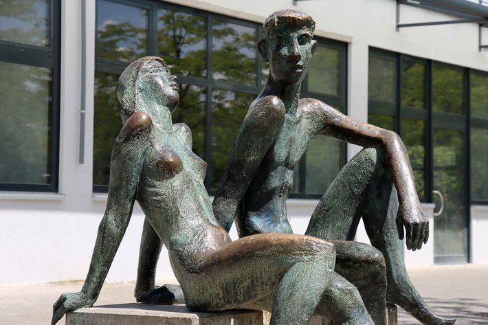 Bronzestatue zeigt zwei sitzende Figuren, eine Frau mit langem Haar, die sich an einen sitzenden Mann mit muskulösem Oberkörper lehnt, beide wirken in inniger Umarmung.