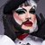 Person mit auffälligem, theatralischem Make-up, darunter dramatische Augenpartie mit schwarzem und blauem Eyeliner, roter Lippenkontur und schwarzem Bart. Tragt einen schwarzen Hut mit Spitzen und ein weißes, spitzenbesetztes Oberteil.