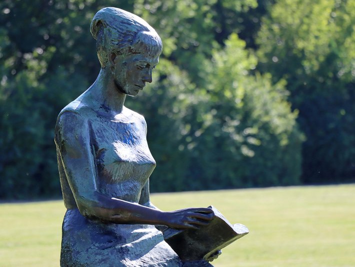 Bronzefigur einer sitzenden Frau, die ein offenes Buch auf dem Schoß hält und nachdenklich in die Ferne blickt.