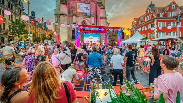 Belebte Straßenveranstaltung mit vielen Menschen, die auf einer Bühne mit bunten Lichtern und Dekorationen zuschauen. Familien, Kinder und Erwachsene genießen ein Fest mit Musik, Ständen und festlicher Atmosphäre bei Sonnenuntergang.