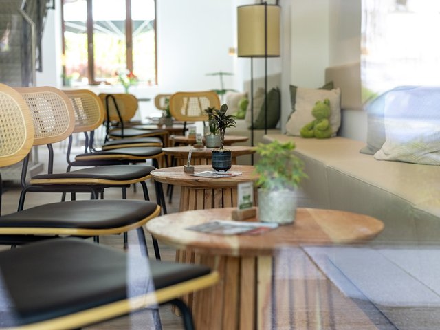 Moderne Café-Ecke mit mehreren schwarzen Stühlen und runden Holztischen, auf denen Gläser, eine Vase mit Pflanze und ein Buch liegen. Im Hintergrund große Fenster mit Tageslicht und eine gemütliche Sitzgruppe mit Kissen.