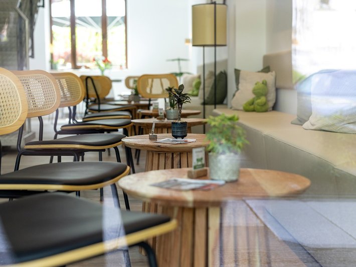 Moderne Café-Ecke mit mehreren schwarzen Stühlen und runden Holztischen, auf denen Gläser, eine Vase mit Pflanze und ein Buch liegen. Im Hintergrund große Fenster mit Tageslicht und eine gemütliche Sitzgruppe mit Kissen.