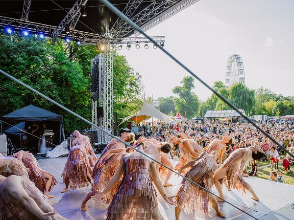 Große Gruppe von Menschen in Kostümen, die auf einer Freilichtbühne vor Publikum tanzen oder performen. Die Kostüme bestehen aus braunen, pelzartigen Materialien, die an Tierfelle erinnern. Im Hintergrund ist eine Bühne mit Technik und ein großer Turm zu sehen.