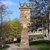 Denkmal mit Uhrenturm und Krone auf dem Dach, umgeben von einem gepflegten Platz mit Bäumen und Gebäuden im Hintergrund, vermutlich historisches oder städtisches Wahrzeichen.