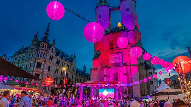 Abendliche Festlichkeitsbeleuchtung mit vielen pink und lila schwebenden Laternen über einer beleuchteten historischen Gebäude-Fassade, vor der sich ein belebter Markt oder eine Veranstaltung mit Menschen und Ständen befindet.