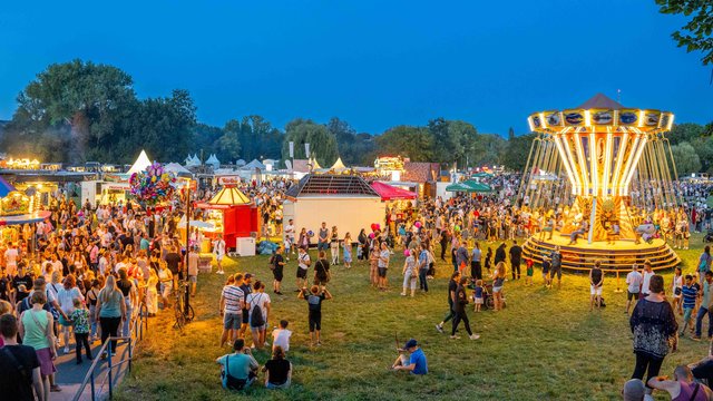 Abendliches Volksfest mit vielen Besuchern auf einer Wiese, beleuchteten Fahrgeschäften wie einem Riesenrad und bunten Ständen, fröhliche Atmosphäre bei Dunkelheit.