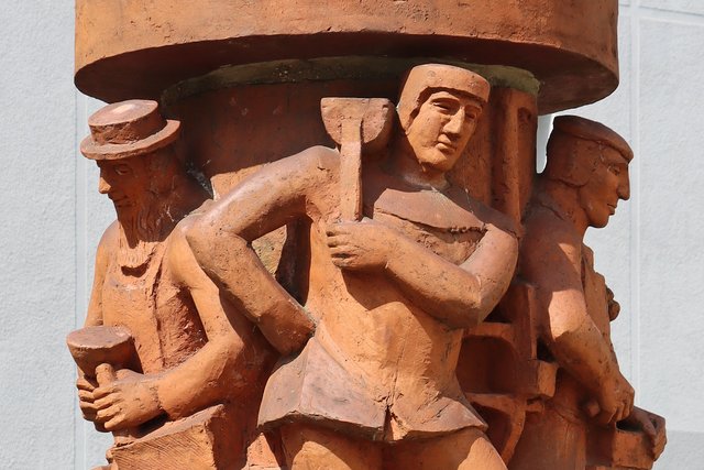 Steinrelief mit drei mittelalterlichen Figuren: Eine Person mit Umhang und Hut steht unter einem Baldachin, während zwei weitere Personen, eine davon mit einem Beutel, scheinbar eine Szene mit einem Stab oder Werkzeug darstellen.