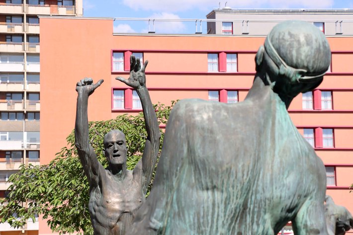 Bronzestatue einer Person, die einen Basketball hält und in einer dynamischen Pose mit ausgestrecktem Arm und winkender Geste steht, vor einem Wohngebäude mit Fenstern und Bäumen.