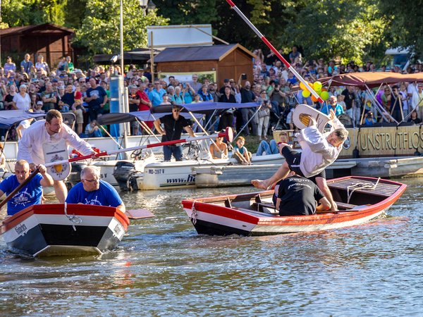 Zwei kleine Boote mit je zwei Personen, die auf einem Fluss fahren, während im Hintergrund eine große Menschenmenge am Ufer steht und zusieht. Die Szene wirkt fröhlich und findet bei gutem Wetter statt.