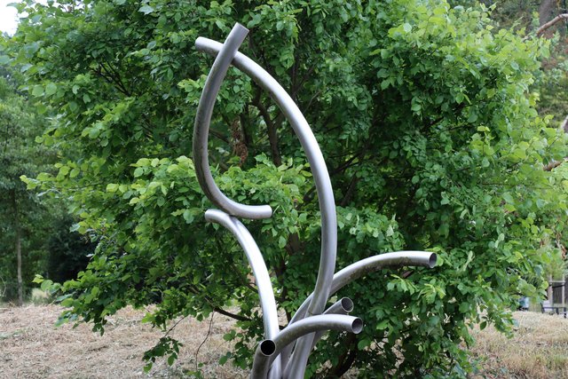 Abstrakte Metallskulptur in Form von geschwungenen, organisch anmutenden Linien, die wie Äste oder Ranken aussehen und sich nach oben verjüngen, umgeben von grünem Blattwerk.