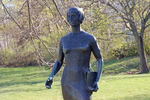 Bronzefigur einer Frau in eleganter Kleidung mit Hut, die eine Handtasche hält und eine Hand an die Hüfte stützt, vor grünem Rasen und Bäumen im Hintergrund.