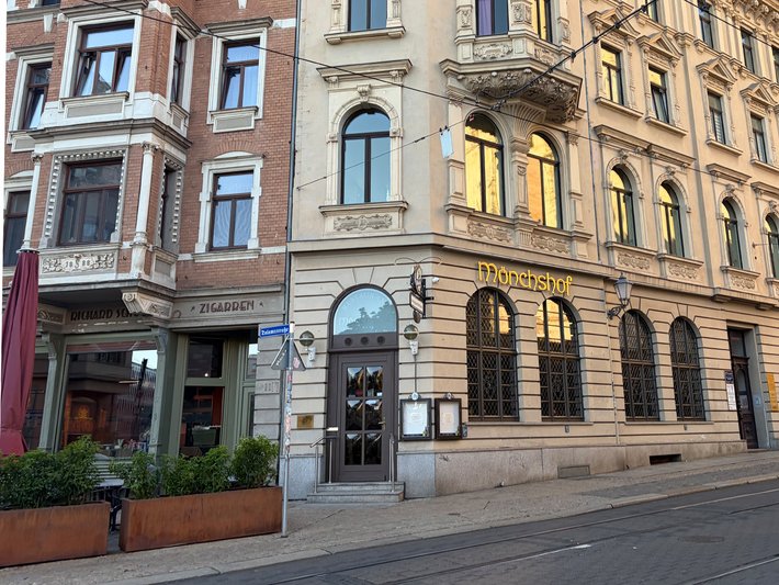 Altbau mit drei Stockwerken und klassizistischer Fassade, Eingangsbereich mit Glas- und Holztüren, davor ein kleiner Außenbereich mit Tischen und Stühlen, vermutlich ein Restaurant oder Café in einer Stadtstraße.