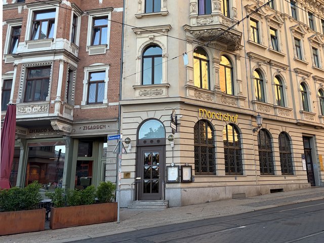 Altbau mit drei Stockwerken und klassizistischer Fassade, Eingangsbereich mit Glas- und Holztüren, davor ein kleiner Außenbereich mit Tischen und Stühlen, vermutlich ein Restaurant oder Café in einer Stadtstraße.