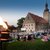 Abendveranstaltung im Freien mit vielen Menschen, die auf Decken und Sitzgelegenheiten sitzen und einem historischen Gebäude mit Turm im Hintergrund, beleuchtet durch eine große Flamme oder Lichtquelle links im Bild.