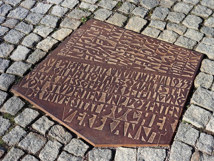 Ein taktiles Bodenleitsystem aus Metall mit erhabenen Braille-Zeichen und Pfeilen, das blinden Menschen als Wegweiser dient, vermutlich für eine bestimmte Richtung oder einen Zielort.