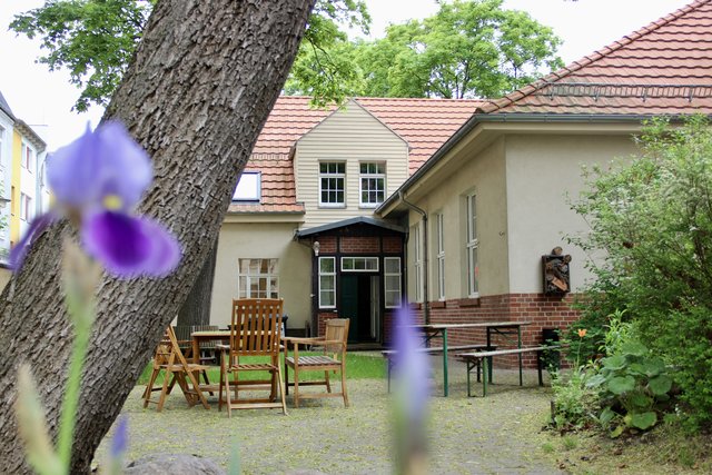 Ein helles, zweistöckiges Haus mit rotem Dach und weißer Fassade, umgeben von einem kleinen Garten. Vor dem Haus stehen mehrere Tische mit Stühlen auf einer Terrasse. Im Vordergrund blühen lila Blüten an einem Strauch.