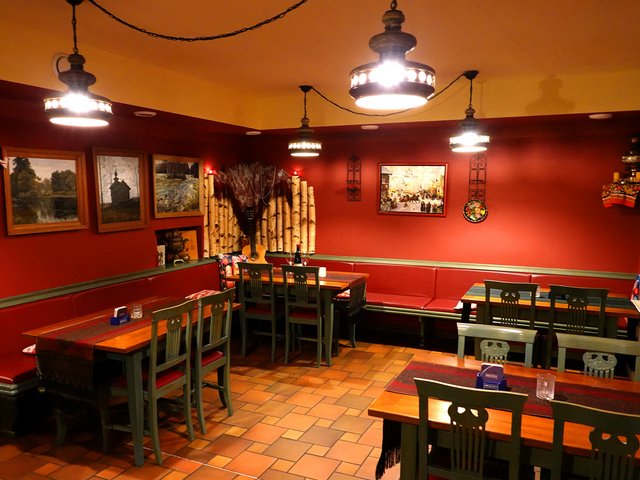 Gemütlicher Raum mit mehreren Holzbänken und Tischen in einer rustikalen Bar oder Gaststätte, rot getönte Wände, warmes Licht und dekorative Elemente wie Bilder und Haken an der Wand.