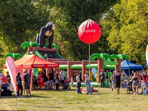 Bunte Familienveranstaltung auf einer Wiese mit Spielbereich, Zeltständen und Menschen, darunter ein großer roter Ballon und ein Spielgerät in Form eines Elefanten.