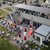 Große Außenveranstaltung mit vielen Gästen, die an Tischen mit roten Sitzgelegenheiten sitzen und sich auf einer Terrasse vor einem Gebäude versammeln. Im Hintergrund sind weitere Menschen auf einer Dachterrasse zu sehen.