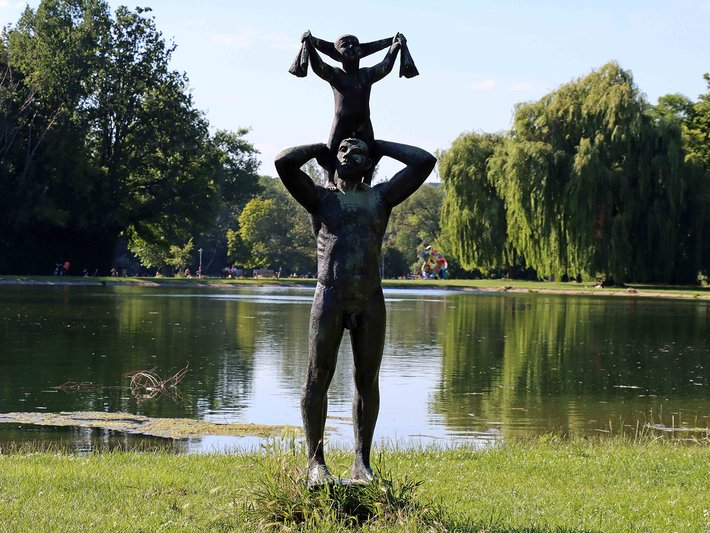 Bronzestatue einer Person, die eine zweite, kleinere Figur auf den Schultern trägt, vor einem ruhigen See mit Gras und Bäumen im Hintergrund.