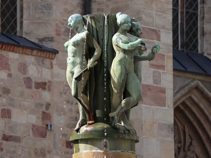 Bronzestatue mit zwei nackten, sich umarmenden Figuren, eine sitzend, eine kniend, die eine Art tragenden oder stützenden Bewegung ausführen, auf einem Sockel vor einem historischen Backsteingebäude.