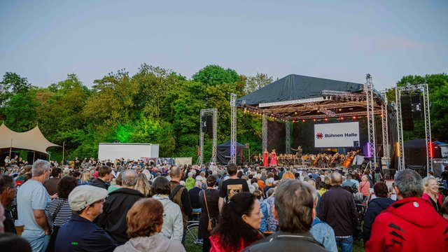 Anna Kolata Großes Open-Air-Konzert mit vielen Zuschauern auf einer Freilichtbühne, die vor einem Wald liegt. Die Bühne zeigt eine Band oder Künstler, die auf der Bühne spielen.