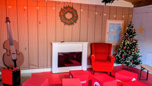 Weihnachtszimmer mit Kamin, rotem Sessel, geschmücktem Weihnachtsbaum, Gitarre an der Wand und festlichem Wandschmuck wie einem Kranz über dem Kamin.