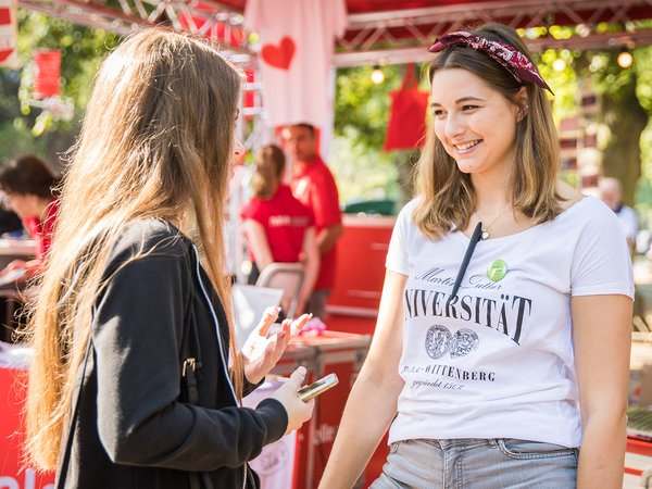 Zwei junge Frauen unterhalten sich lächelnd auf einer Veranstaltung, wobei eine ein weißes T-Shirt mit dem Aufdruck „Universität“ und „Baden-Württemberg“ sowie eine grüne Schleife trägt, während die andere ein dunkles Oberteil mit Kapuze und eine Brille mit blauer Fassung hat.