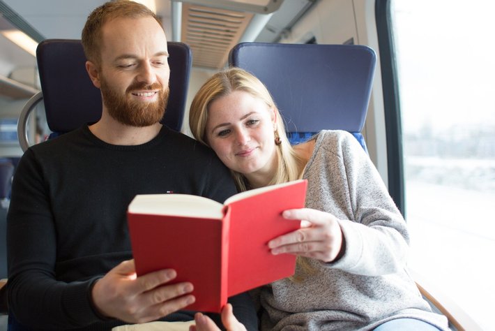 Zwei Personen auf einem Zug sitzen gemeinsam eng beieinander und blättern gemeinsam in einem roten Buch. Die Frau zeigt auf eine Seite, der Mann blickt aufmerksam in das Buch.