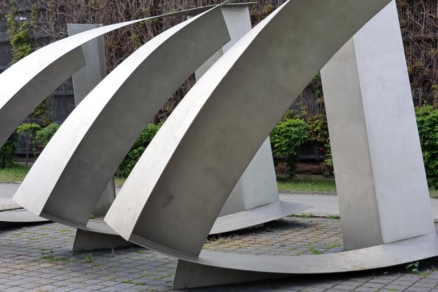 Drei große, stählerne, wellenförmige Skulpturen, die sich nach oben verjüngen und wie schwebende Bögen wirken, angeordnet in einer Reihe auf einem gepflasterten Platz.