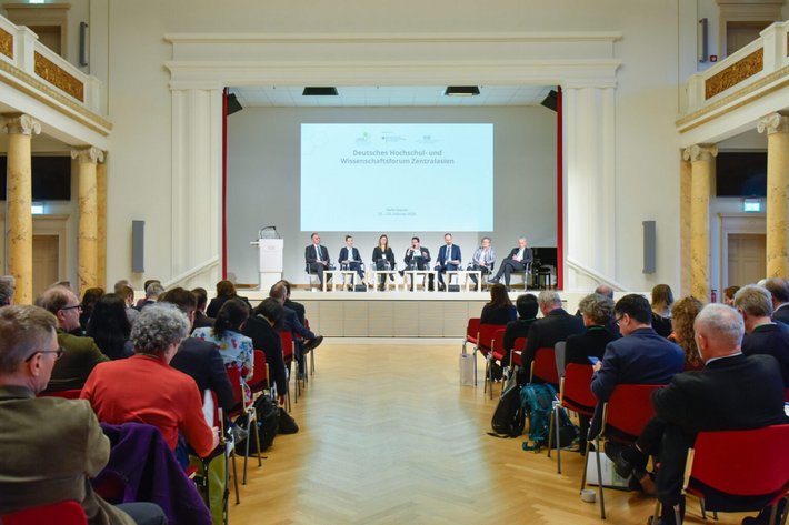 Großer Saal mit Publikum, das auf roten Stühlen einer Veranstaltung beiwohnt. Auf der Bühne stehen mehrere Personen, vermutlich Redner oder Moderatoren, vor einem großen Bildschirm mit Präsentation.