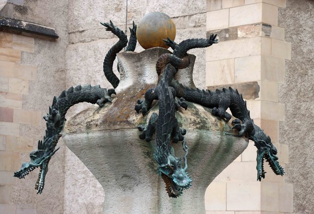 Bronzene Skulptur eines Drachen mit weit aufgerissenem Maul, der sich um einen runden Gegenstand wie eine Kugel windet, auf einem steinernen Sockel.