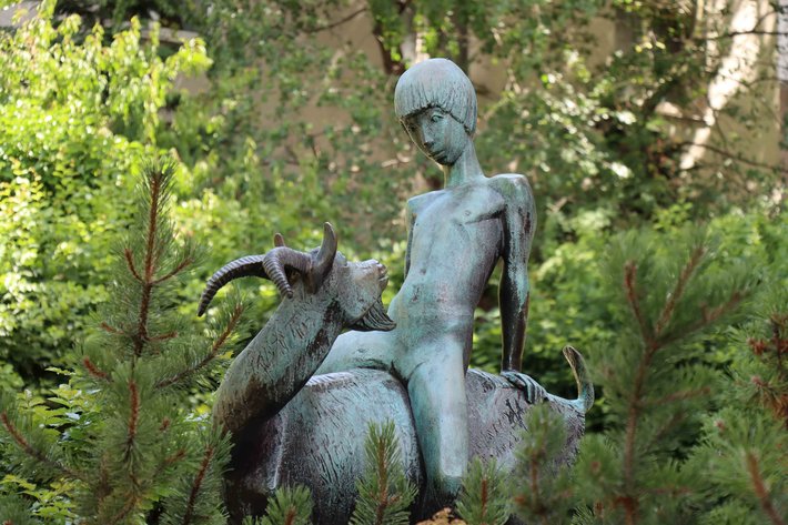 Bronzefigur eines Mädchens, das auf einem großen, stilisierten Fisch reitet, umgeben von dichter Natur mit Bäumen und Büschen im Hintergrund.