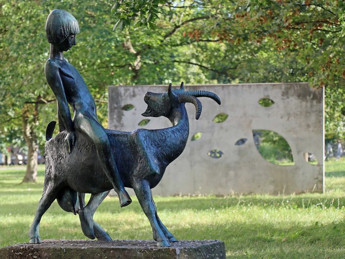 Bronzefigur eines Menschen, der auf einem großen, stilisierten Vogel reitet, der wie ein Huhn oder eine Ente aussieht, im Freien aufgestellt.