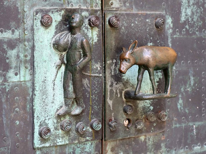 Altar mit Reliefdarstellung einer Person, die ein Kind auf dem Arm hält, daneben ein Relief eines Rehs, beide Figuren in metallischer Ausführung an einer alten Tür.