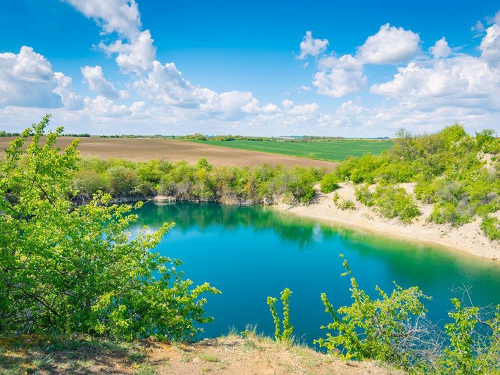 Klare, tiefblaue Wasserfläche inmitten einer grünen, leicht hügeligen Landschaft mit Büschen und Bäumen am Ufer, sonniges Wetter mit blauen Himmel und weißen Wolken.