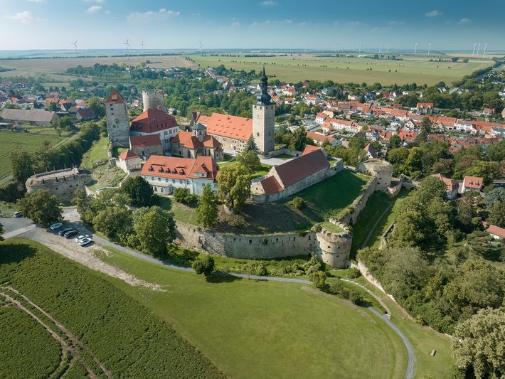 Luftbild einer historischen Burg mit roten Dächern und weißer Mauer, umgeben von grünen Wiesen und Feldern, im Hintergrund eine kleine Stadt mit Kirche und Häuserzeilen.
