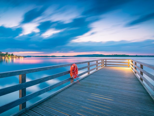 Holzstege führt über ruhiges Gewässer bei Dämmerung, am Ende ein rotes Rettungsring-Symbol an der Reling, sanfte Abendstimmung mit spiegelndem Wasser und bläulichem Himmel.