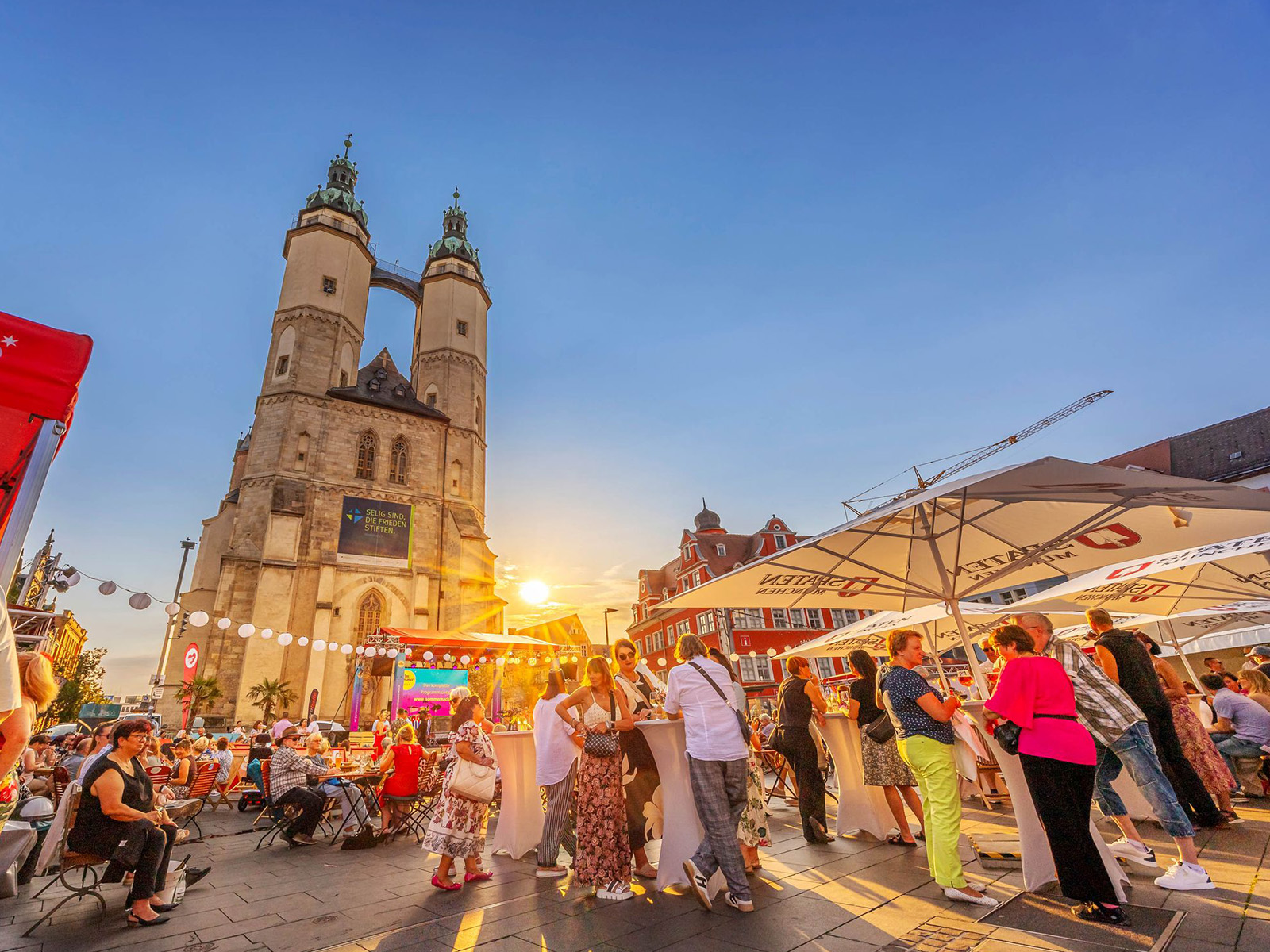 Abendliche Markt- oder Festplatzszene mit beleuchteter historischer Kirche im Hintergrund, zwei markanten Türmen und Kuppel. Vorne viele Menschen, die an Ständen mit bunten Markisen essen, trinken und sich unterhalten. Lebhafte Atmosphäre bei Sonnenuntergang.
