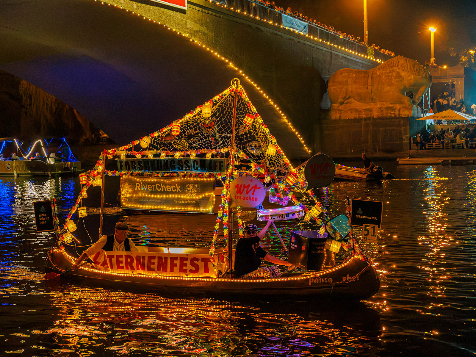 Bootsfestlich geschmücktes Boot auf dem Wasser während des Laternenfestes, beleuchtet mit vielen bunten Lichterketten und Dekorationen, im Hintergrund eine beleuchtete Brücke.