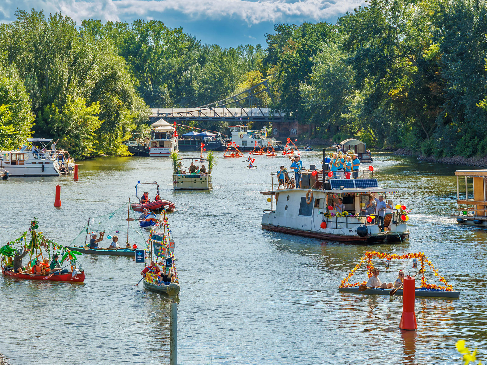 Bunte Bootsverleih-Station am Flussufer mit vielen gemieteten Tretbooten auf dem Wasser, die von Menschen genutzt werden, umgeben von grüner Landschaft und einer Brücke im Hintergrund.