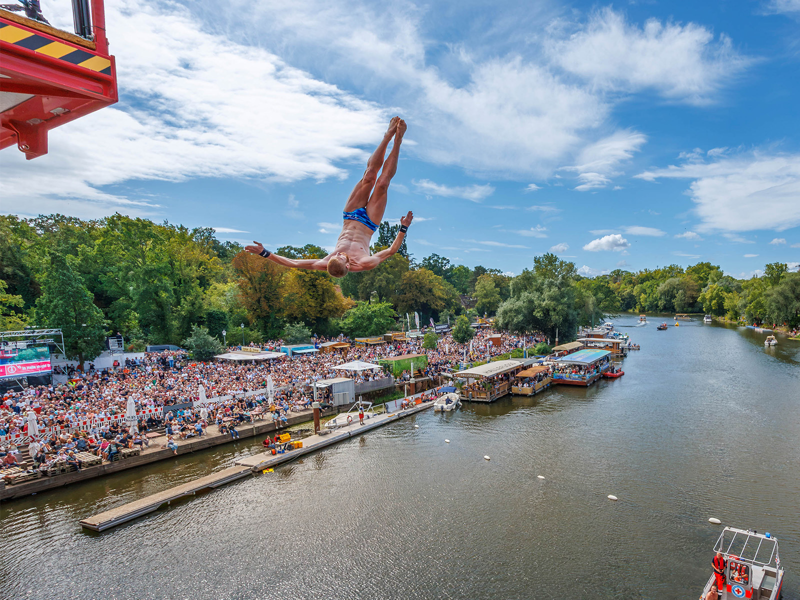 Großer Kran hebt im Hintergrund einen Lastwagen oder Container über einem belebten Flussufer mit vielen Menschen, die ein Fest oder eine Veranstaltung besuchen. Im Vordergrund sind Boote auf dem Wasser zu sehen.