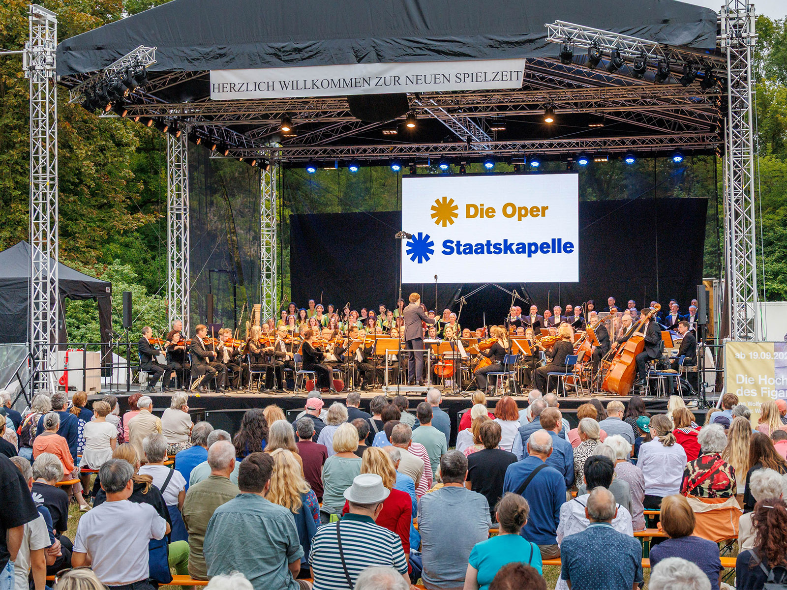 Großes Orchester der Staatskapelle auf einer Freilichtbühne mit dem Schriftzug „Die Oper“ spielt vor Publikum im Freien.