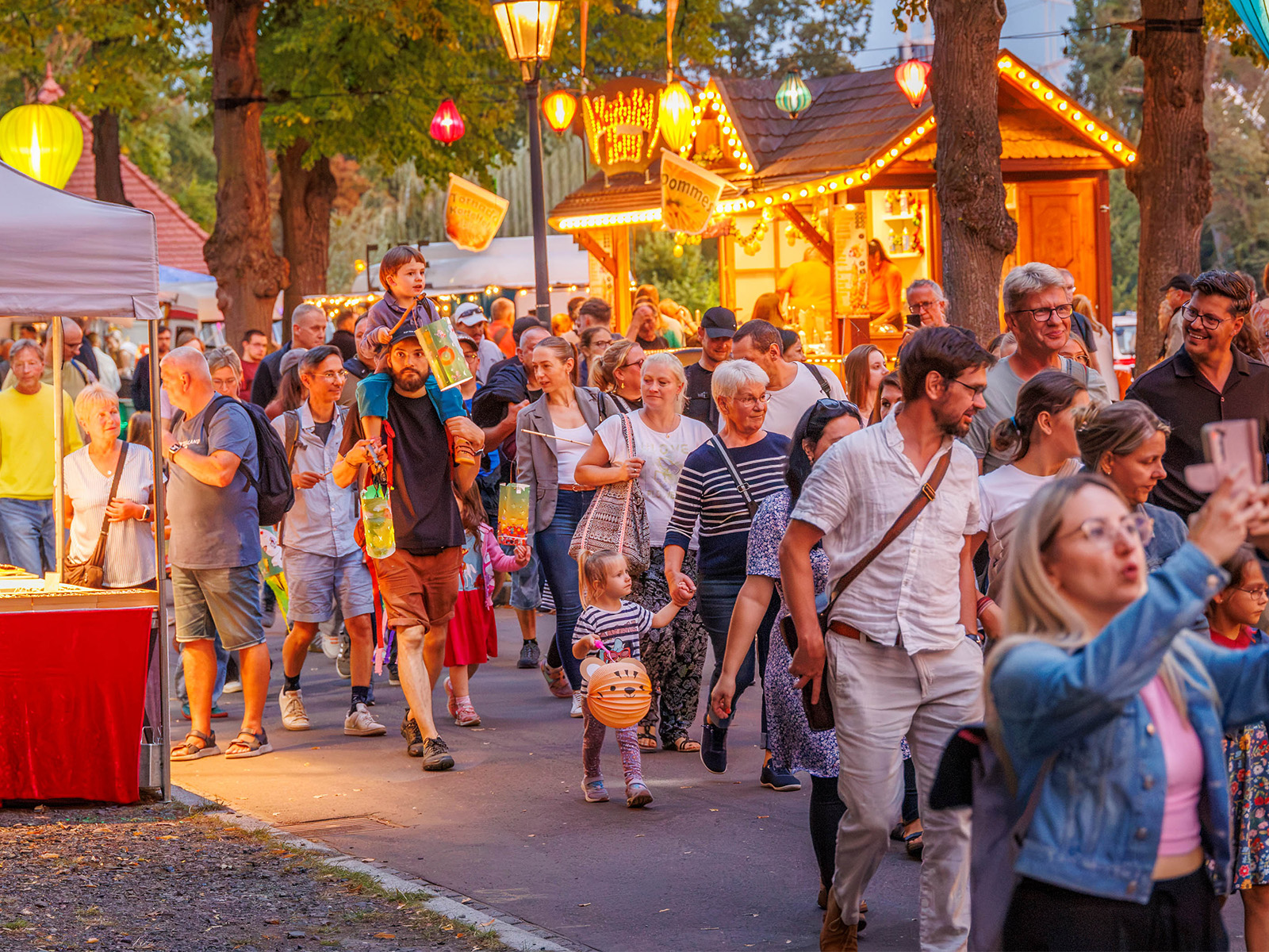 Belebte Abendstraße mit vielen Menschen, die spazieren, plaudern und sich unterhalten. Im Hintergrund leuchtet ein Marktstand mit warmem Licht, daneben ein Restaurant mit Terrasse und Laternen. Gemütliche Atmosphäre bei Dämmerung.